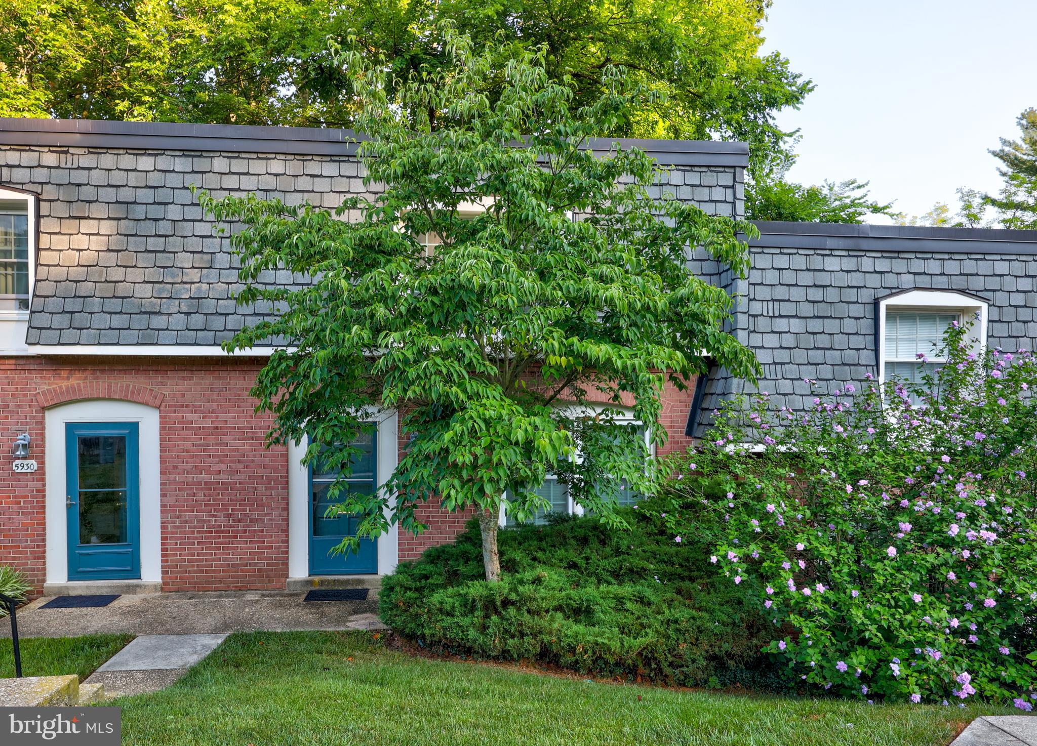 a view of a brick house with a yard plants and large tree