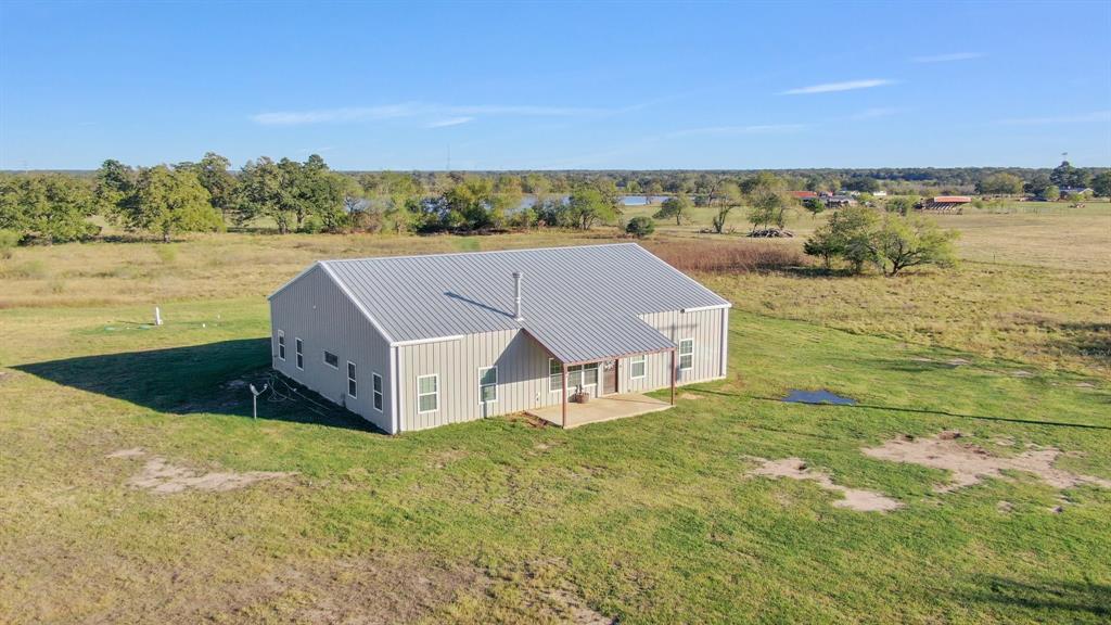 This is a modern, single-story home with a metal roof, set in a spacious rural setting with open grassland. The house has a covered front porch and appears to be in a tranquil area, offering a sense of privacy and open space.