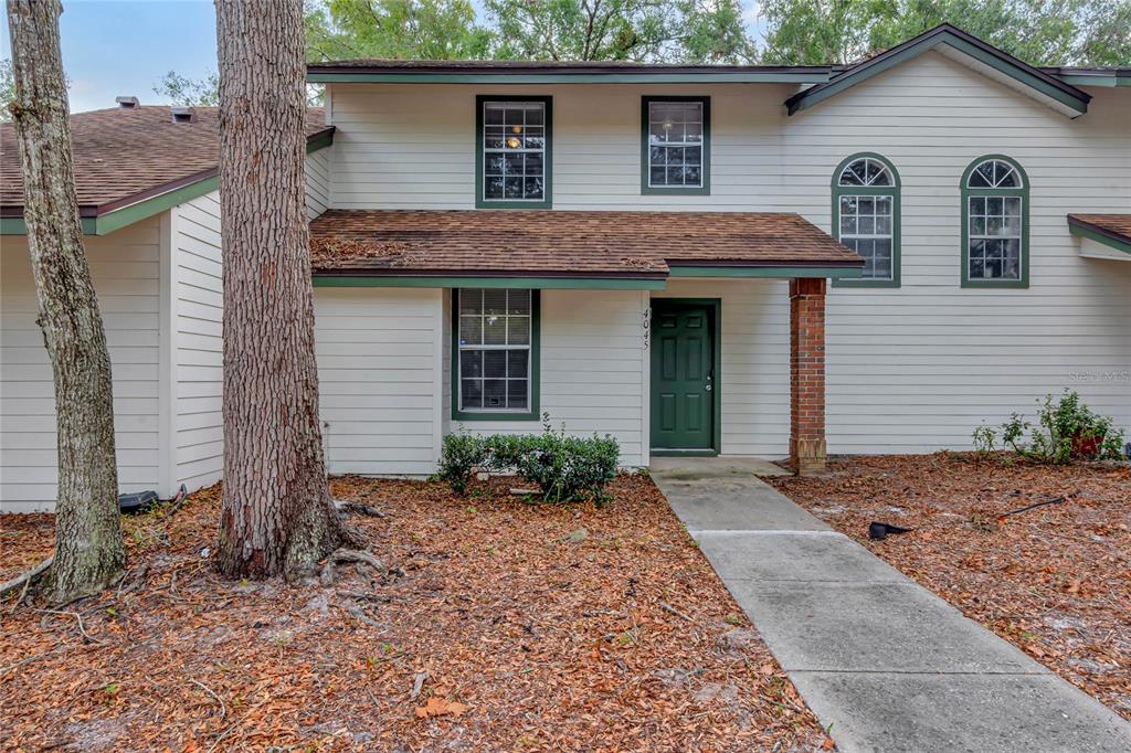 The image shows the exterior of a two-story townhouse with white siding and a brown shingled roof. The entrance is centered, featuring a green door with a small concrete pathway leading up to it. The windows on the second floor have a distinct design, with two tall, arched windows on the right and rectangular windows on either side of the front door. A large tree with a thick trunk is on the left side of the yard, which is covered in dry leaves and mulch with some low green shrubs near the house. The adjacent unit, partially visible on the right, has a similar design, and an ADT security sign is placed near its entrance. The sky is blue, and the area is surrounded by leafy trees.