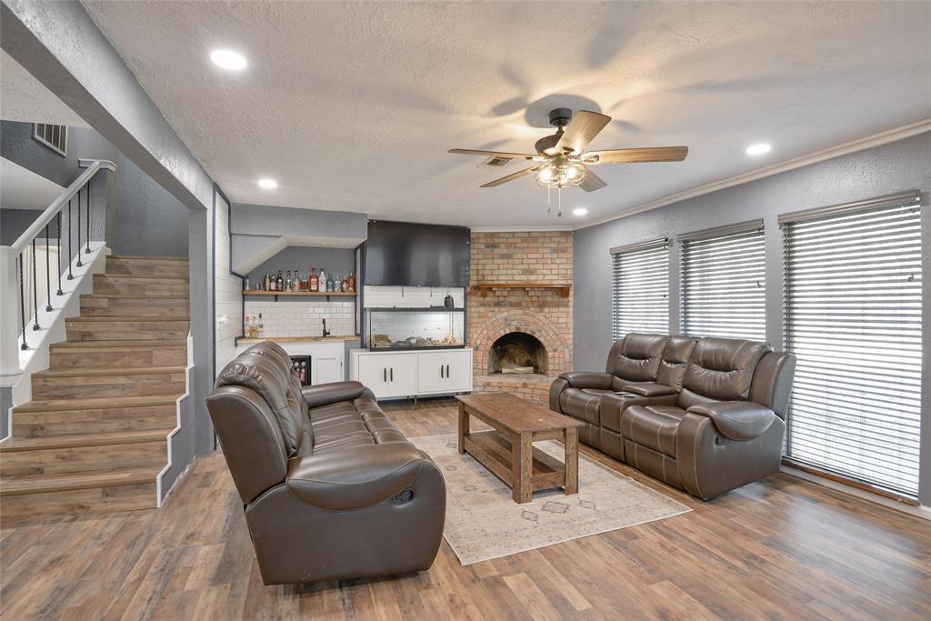 This is a spacious, open-plan living area featuring wood flooring, a cozy brick fireplace, modern ceiling fan, and a staircase leading to the upper level. The room is well-lit with natural light from large windows and includes a comfortable seating area. An adjacent kitchen with a white countertop and bar seating can also be seen.