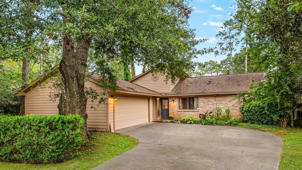 This is a single-story home with a beige exterior, featuring a two-car garage and a large driveway. The house is nestled among mature trees and greenery, providing a serene and shaded setting.