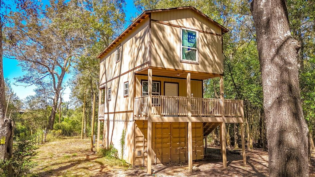 This is a two-story wood-sided home elevated on stilts, featuring a spacious balcony, surrounded by a natural wooded setting.