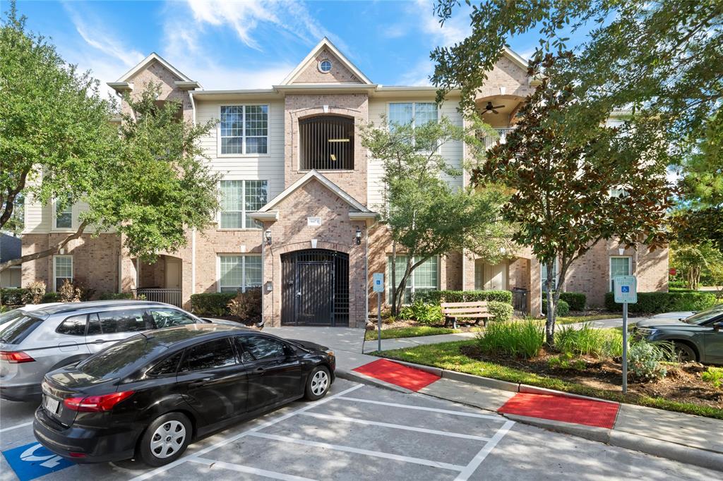 This photo shows a multi-story residential building with brick facade, large windows, and a gated entrance, surrounded by mature trees and well-maintained landscaping. There is parking available in front of the building.