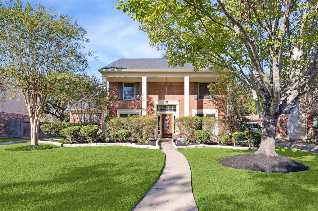 Charming two-story brick home with well-manicured lawn and mature trees, offering a welcoming walkway.