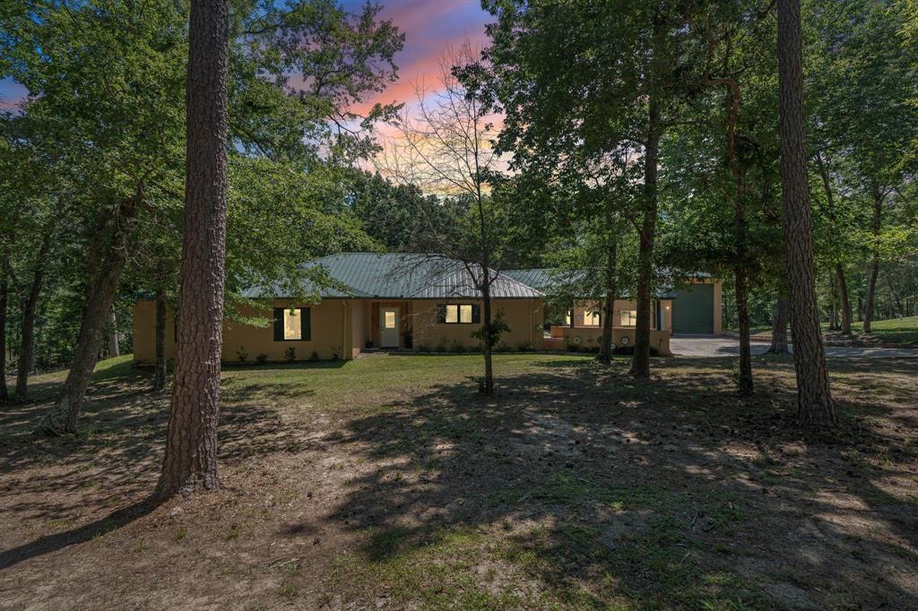 Welcome home, this beautiful green metal roof home is nestled among tall trees and greenery. A large garage is visible to the right, and the driveway curves around the property, surrounded by a forested area. Gives a feeling of peace and seclusion, blending harmoniously with its natural surroundings.