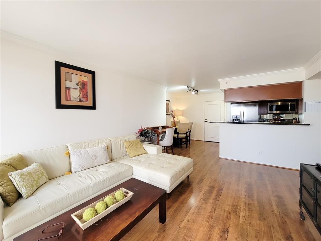 Bright, open-concept living space with a comfortable seating area featuring a sectional sofa and laminate flooring that flows seamlessly into the kitchen with a breakfast bar and modern appliances. The space is well-lit and includes tasteful, minimalistic decor.