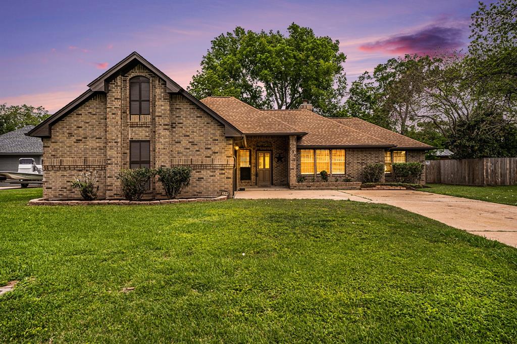 This is a single-story brick home featuring a prominent gable, an attached garage to the left, a large front lawn, and mature trees in the background. The photo shows the house at dusk with interior lights on and a tranquil sky above.