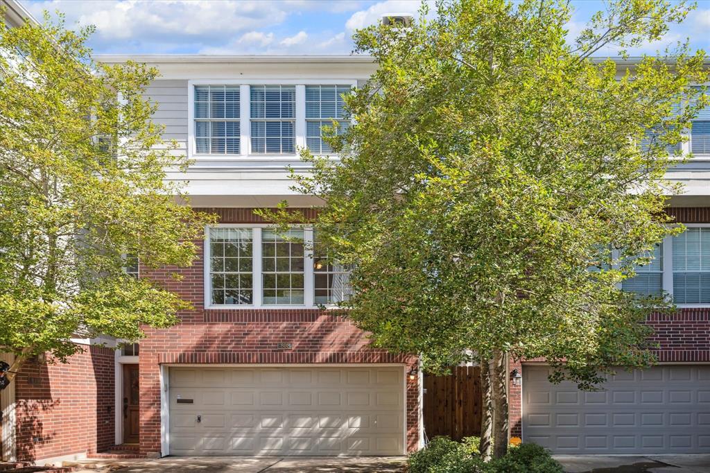 This three-story townhome features a brick exterior, a two-car garage, and mature trees in the front, providing shade and a touch of greenery.