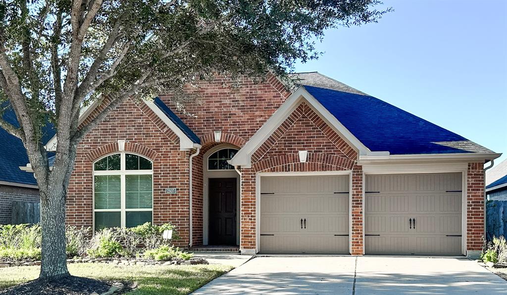 This is a single-story brick home with a prominent arched entryway and a two-car garage. The residence features large windows and is shaded by mature trees.