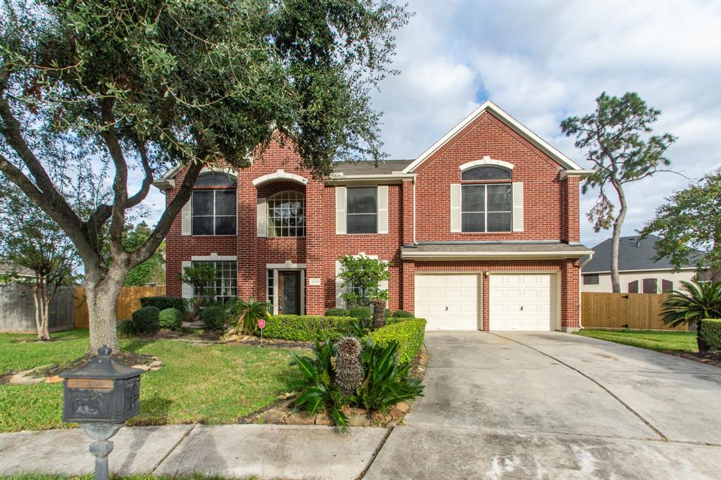 This is a two-story brick house with a double garage, a well-maintained front yard, and mature trees. It features large windows for natural light and a spacious driveway.