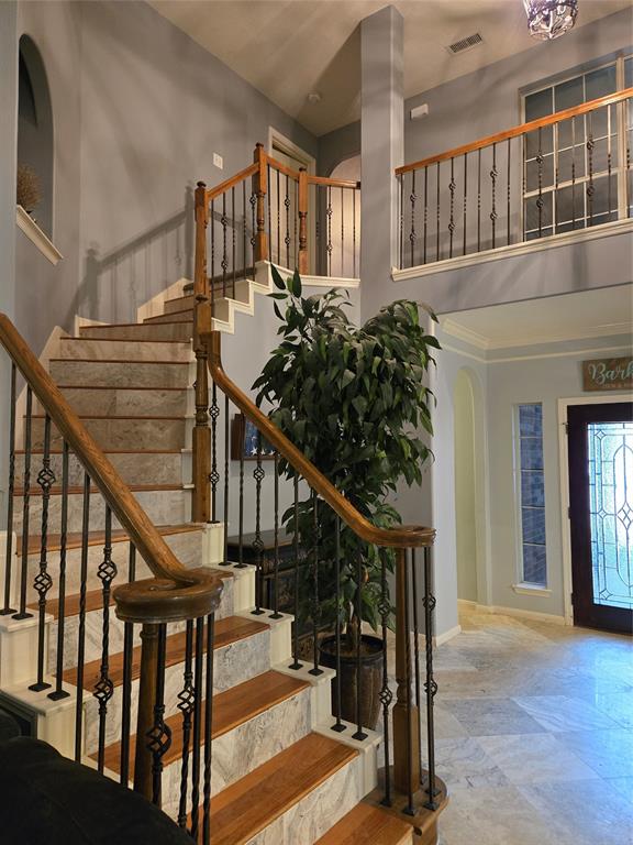 This interior shot showcases an elegant staircase with wood and wrought iron railings, leading to an upper level with a balcony. The space features high ceilings, marble flooring, and neutral walls, which convey a sense of luxury and spaciousness.