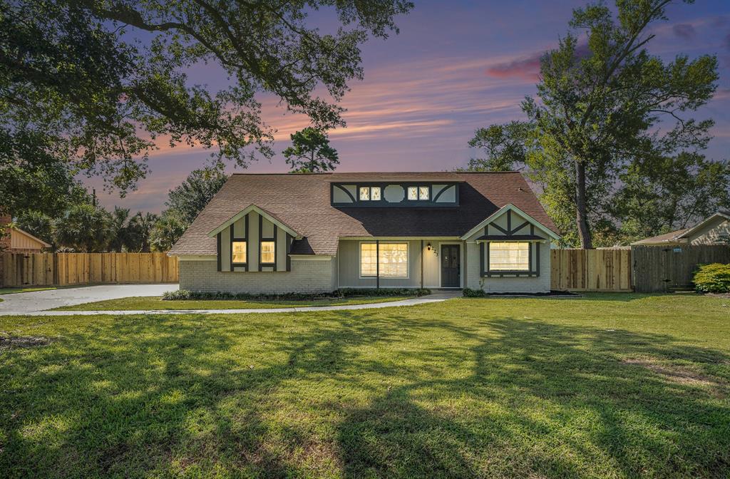 Charming home with a Tudor-style facade, featuring a spacious driveway, large windows, and a backdrop of a beautiful sunset sky. The property includes a well-maintained lawn and privacy fencing.