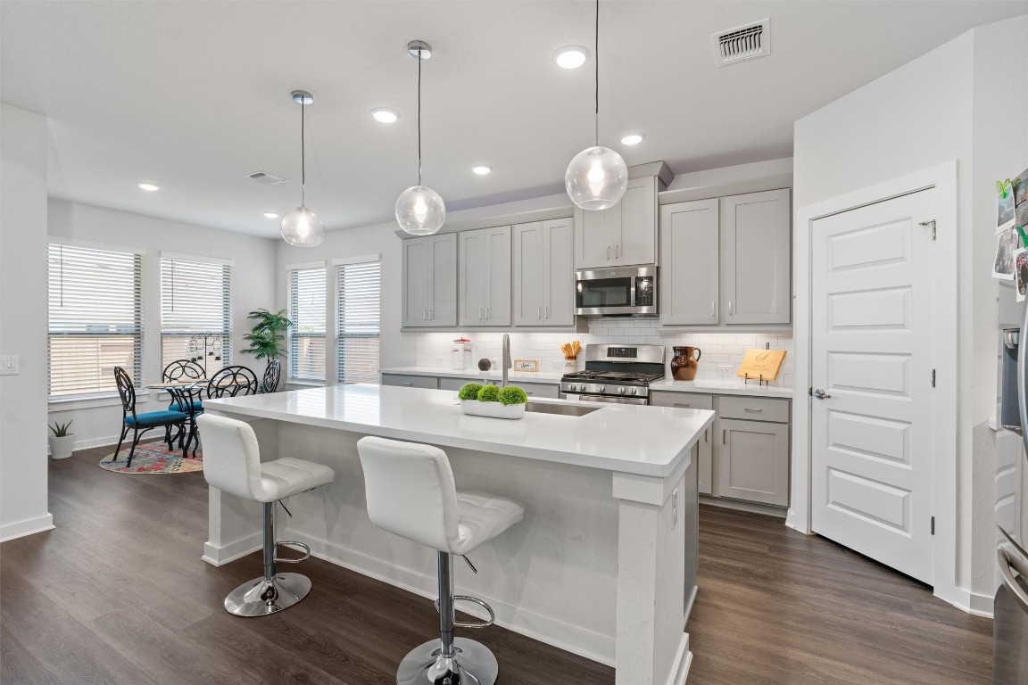 Gourmet kitchen with an expansive quartz center island + sink & filtered water, tastefully accented by modern lighting, subway tile backsplash, a chic neutral color pallet & open to the spacious family room