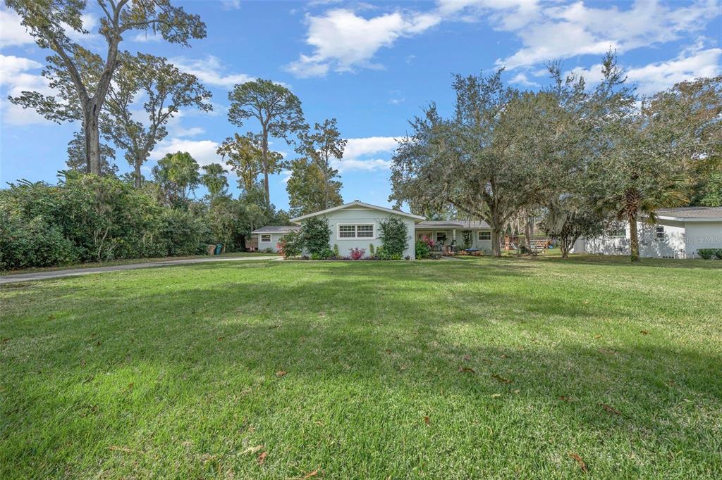 a view of a white house in front of a big yard with plants and large trees