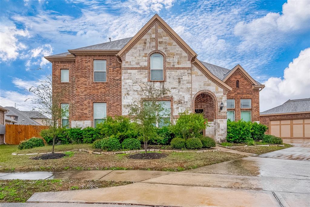 A charming two-story brick and stone house, complete with a front yard and driveway. The home boasts expansive windows and a gracious, arched entryway.