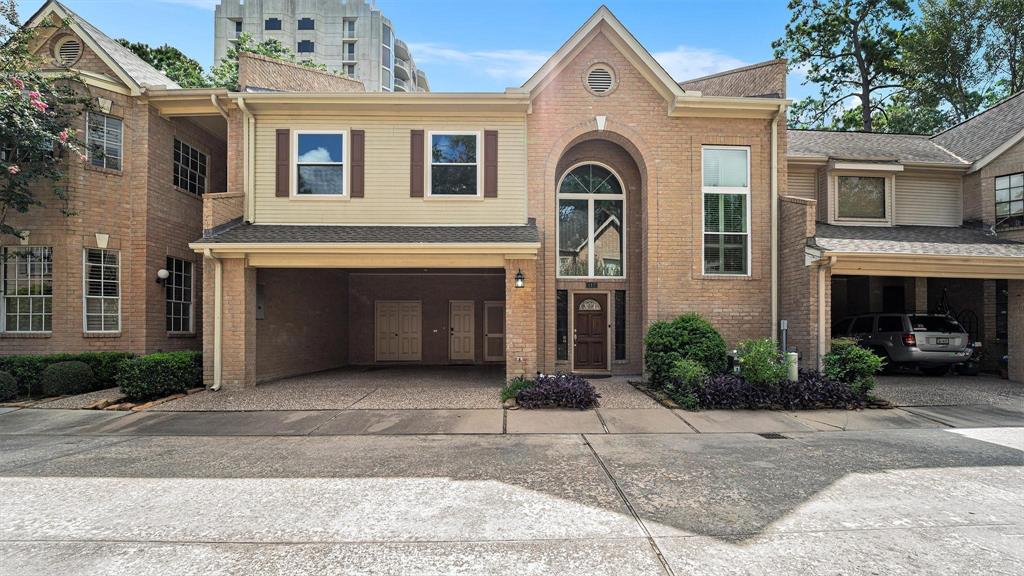 This is a two-story brick townhouse featuring a tall entryway with an arched window, a two-car carport, and contrasting beige siding on the upper level. The home is part of a connected row, nestled in a neighborhood with mature landscaping.