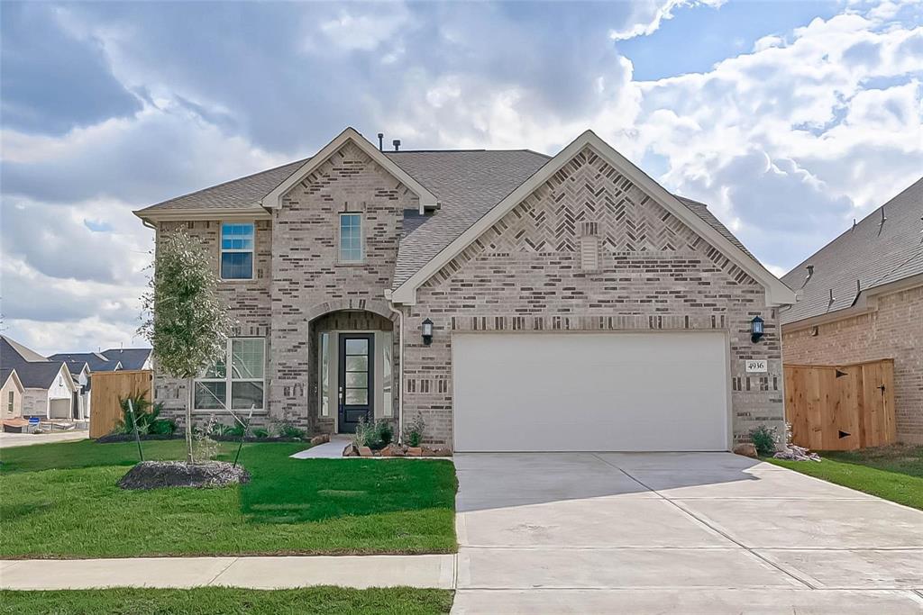 This photo shows a modern two-story home with a brick exterior and a gabled roof. It features a spacious two-car garage and a well-maintained front lawn. The entrance is accented with an arched doorway and the neighborhood appears to be quiet and residential.