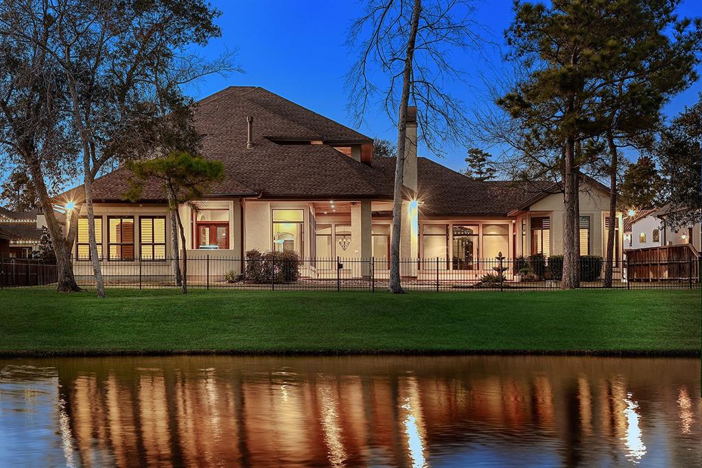 This stunning home features a spacious, elegant design with large windows and beautiful lighting, set against a serene lakeside backdrop. The manicured lawn and trees enhance the inviting atmosphere.