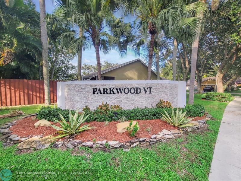 a sign that is sitting in front of a brick house with large tree and wooden fence