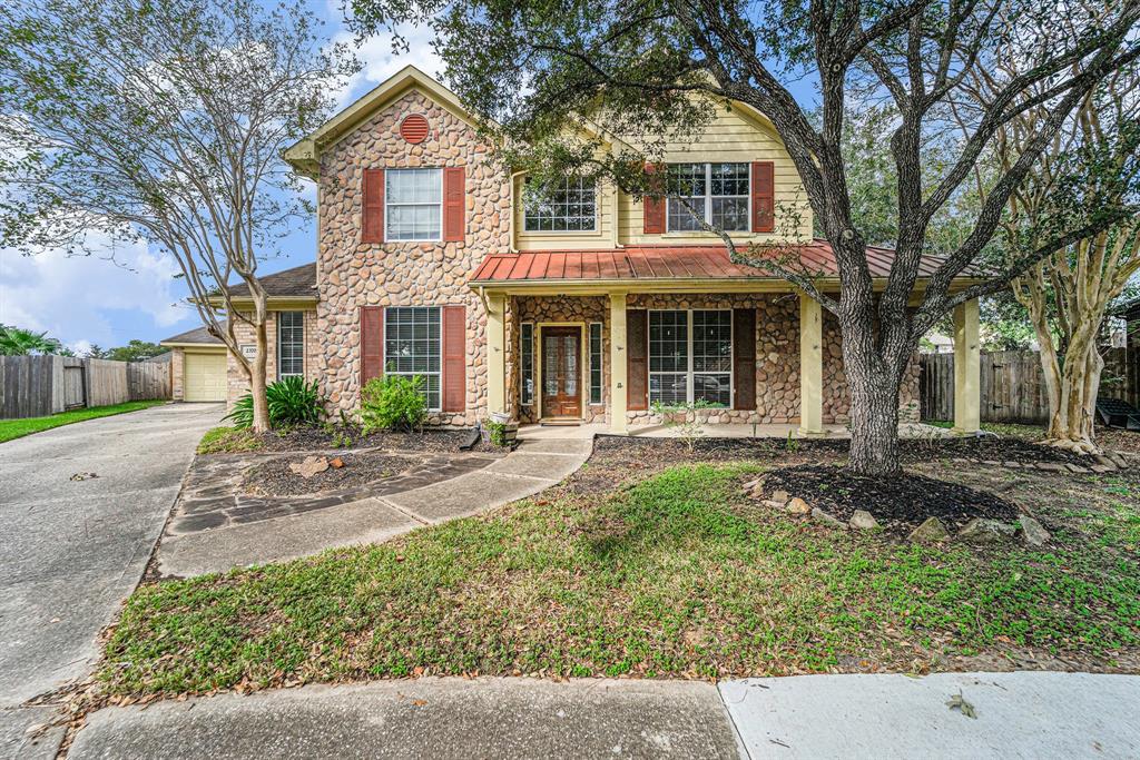 This is a two-story home with a stone and siding exterior, featuring a red tile accent over the second-story window and a matching front door. The house has a single-car garage, a paved driveway, and is shaded by mature trees. It offers a welcoming front porch, nestled within a residential neighborhood.