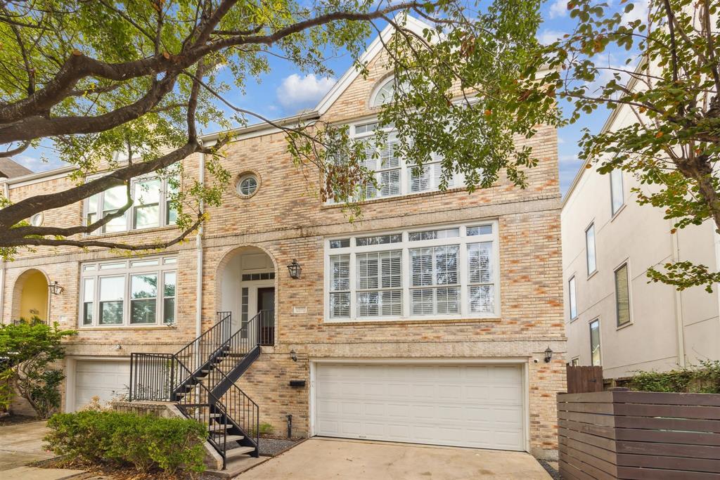 This is a three-story brick townhouse with large windows, featuring a two-car garage and a welcoming entrance up a flight of stairs, nestled among mature trees in a residential neighborhood.