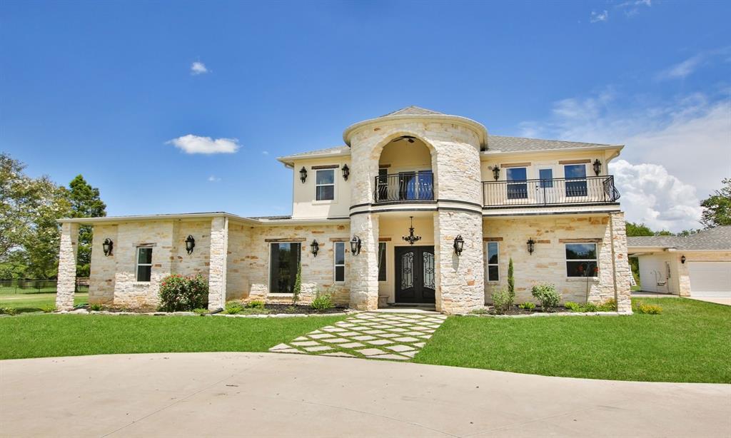 This is a two-story stone facade house featuring a grand entrance with an arched doorway, balcony, and a three-car garage, set against a clear blue sky.