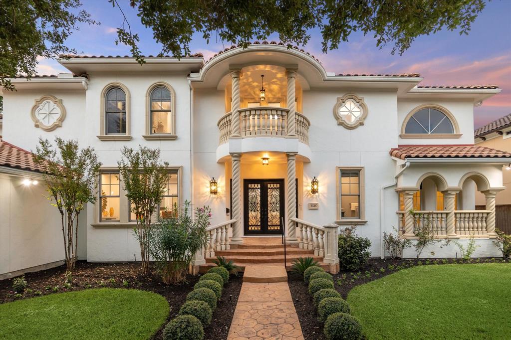 This beautiful Mediterranean-style house features a grand, arched entryway with ornate wrought-iron doors, a balcony with decorative columns, and lush landscaping. The home's elegant architecture, warm lighting, and well-manicured grounds create an inviting and luxurious atmosphere.