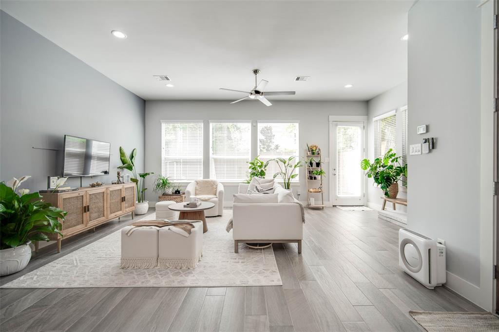 This is a bright, contemporary living room with gray walls, large windows for natural light, wood-look flooring, and modern furnishings. The space feels open and airy, accented with indoor plants and neutral tones.