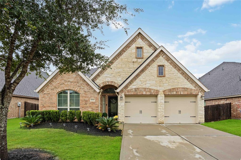 This is a single-story brick 4 Side brick and stone facade home featuring a well-manicured lawn, an arched entryway, and a double garage. 