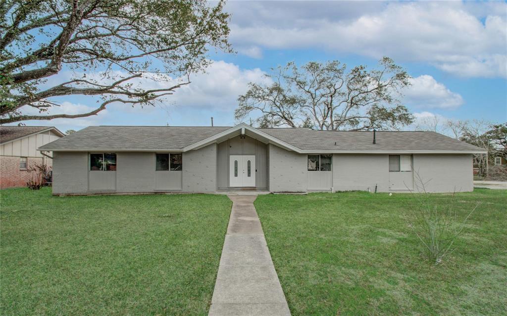 This is a single-story home featuring a light-colored exterior with a well-maintained front lawn and a concrete walkway leading to the central entrance. The house has a simple design with a symmetrical facade, large windows, and is shaded by mature trees.